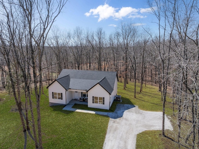 view of front facade with a front yard, a wooded view, dirt driveway, and a shingled roof