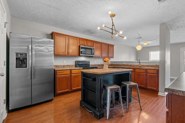 kitchen with wood finished floors, a sink, appliances with stainless steel finishes, brown cabinets, and a chandelier