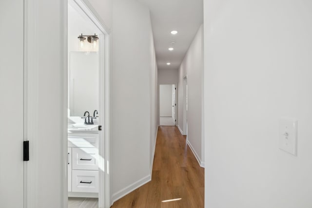 hallway featuring light wood finished floors, recessed lighting, baseboards, and a sink