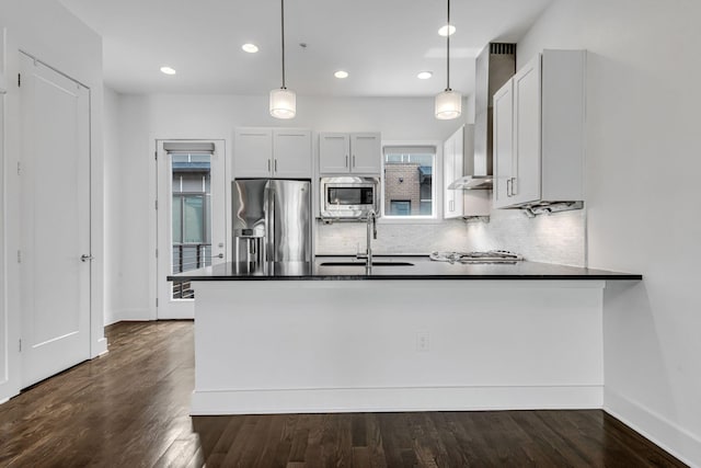 kitchen featuring dark countertops, wall chimney range hood, appliances with stainless steel finishes, a peninsula, and a sink