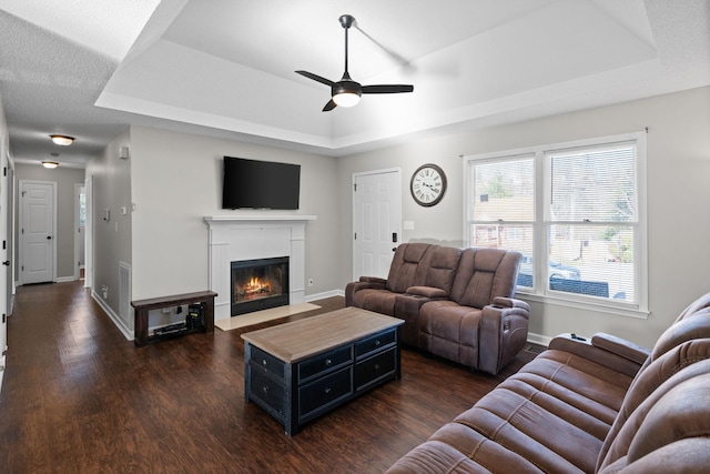 living room with dark wood finished floors, a tray ceiling, a fireplace with flush hearth, and baseboards