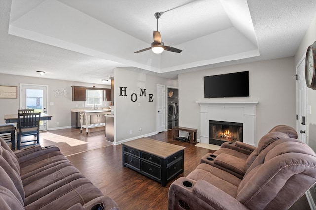 living area with baseboards, a tray ceiling, a fireplace with flush hearth, ceiling fan, and dark wood-type flooring