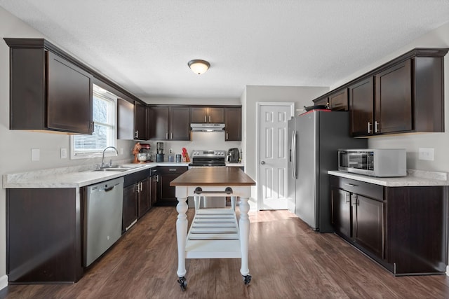 kitchen with under cabinet range hood, dark brown cabinetry, appliances with stainless steel finishes, dark wood-style floors, and a sink