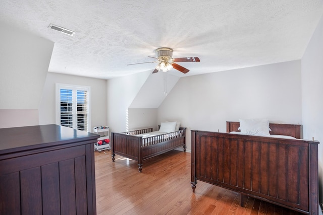 bedroom with light wood finished floors, visible vents, vaulted ceiling, a textured ceiling, and a ceiling fan