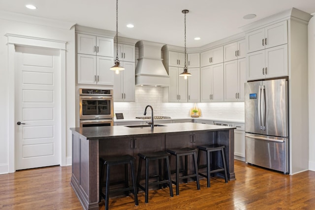 kitchen with dark wood-type flooring, premium range hood, an island with sink, stainless steel appliances, and a sink