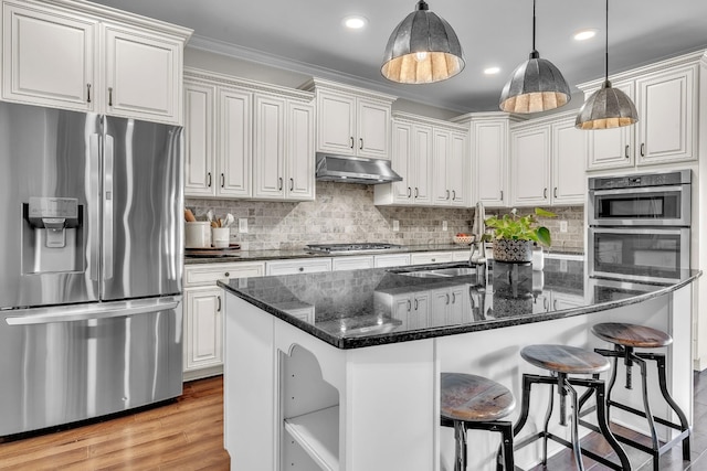 kitchen with light wood finished floors, under cabinet range hood, appliances with stainless steel finishes, crown molding, and backsplash