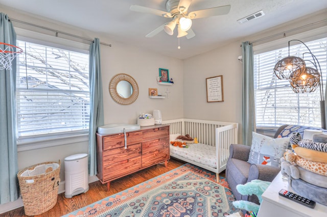 bedroom featuring visible vents, a nursery area, wood finished floors, and a ceiling fan