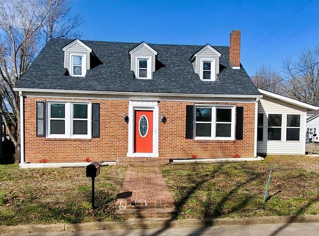 new england style home with brick siding, a chimney, a front yard, and a shingled roof