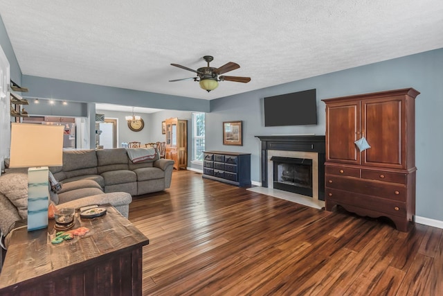 living area featuring a fireplace with flush hearth, dark wood-type flooring, ceiling fan with notable chandelier, a textured ceiling, and baseboards