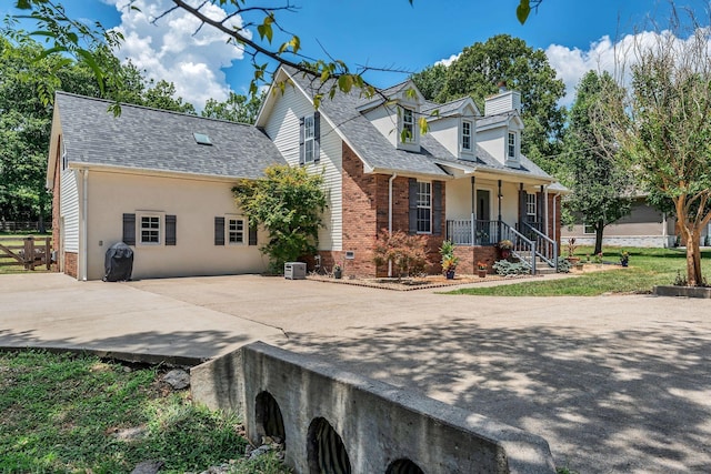 view of front of property with cooling unit, roof with shingles, covered porch, a chimney, and brick siding