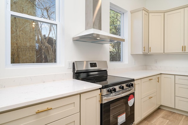 kitchen with wall chimney range hood, light stone countertops, stainless steel electric stove, cream cabinets, and light wood-style floors