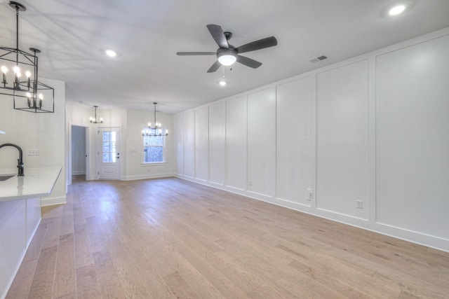 unfurnished living room with visible vents, ceiling fan with notable chandelier, a sink, light wood-style floors, and a decorative wall