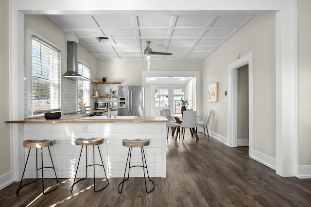 kitchen with baseboards, wooden counters, dark wood-type flooring, wall chimney exhaust hood, and stainless steel fridge