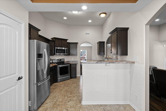 kitchen with light stone counters, a peninsula, arched walkways, stainless steel appliances, and dark brown cabinets