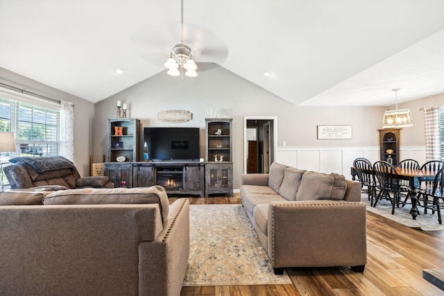 living room featuring a wainscoted wall, lofted ceiling, wood finished floors, a glass covered fireplace, and a ceiling fan