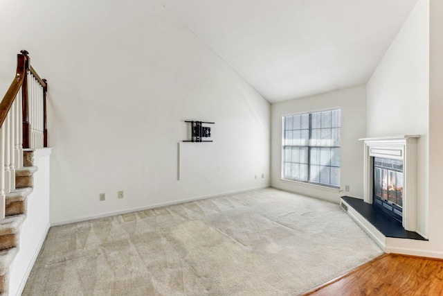 unfurnished living room with high vaulted ceiling, a glass covered fireplace, stairway, carpet floors, and baseboards