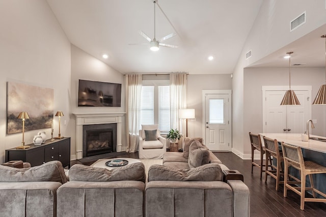 living room featuring a ceiling fan, visible vents, baseboards, dark wood finished floors, and a glass covered fireplace