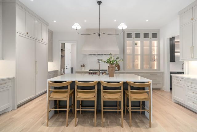 kitchen featuring an island with sink, light wood finished floors, ventilation hood, and light countertops