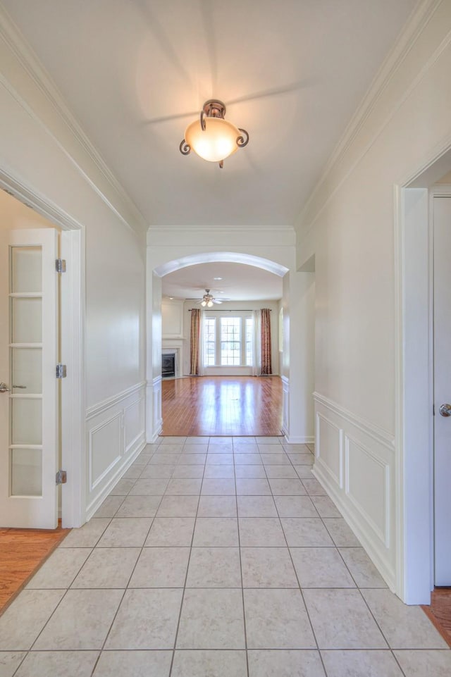 corridor with a wainscoted wall, arched walkways, crown molding, a decorative wall, and light tile patterned floors