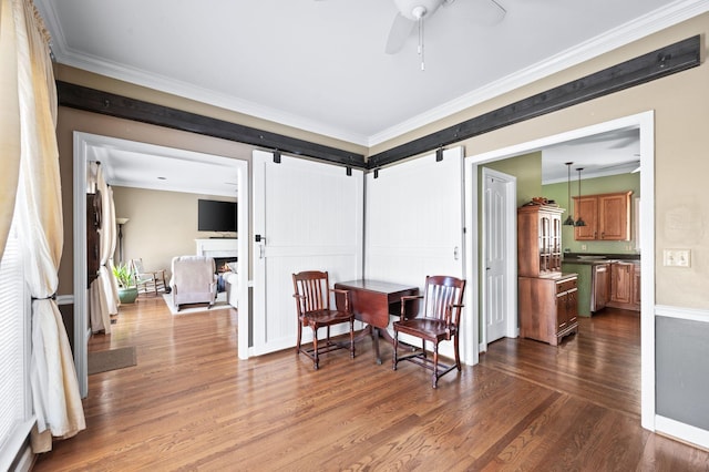 dining space with ornamental molding, a ceiling fan, a warm lit fireplace, dark wood finished floors, and a barn door