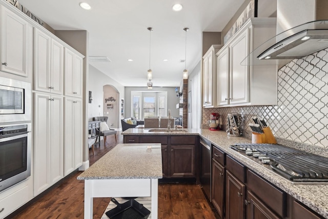 kitchen with arched walkways, a sink, appliances with stainless steel finishes, wall chimney range hood, and backsplash