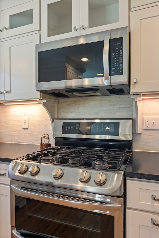 kitchen with tasteful backsplash, dark countertops, white cabinetry, appliances with stainless steel finishes, and glass insert cabinets