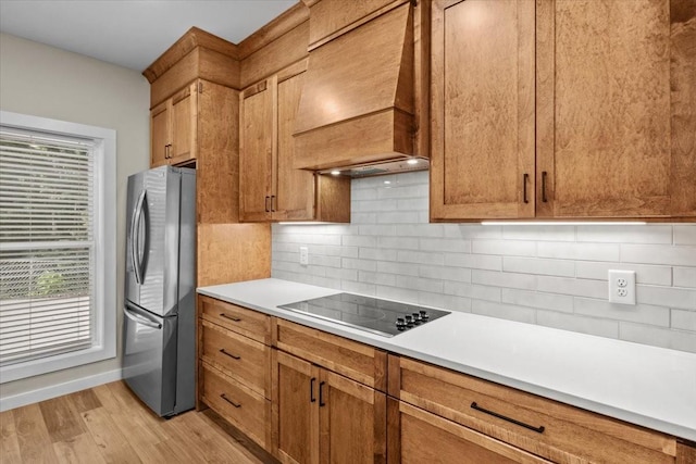 kitchen with freestanding refrigerator, black electric cooktop, light wood-style floors, premium range hood, and decorative backsplash
