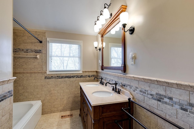 full bathroom featuring a tub to relax in, a wainscoted wall, double vanity, a sink, and tile walls