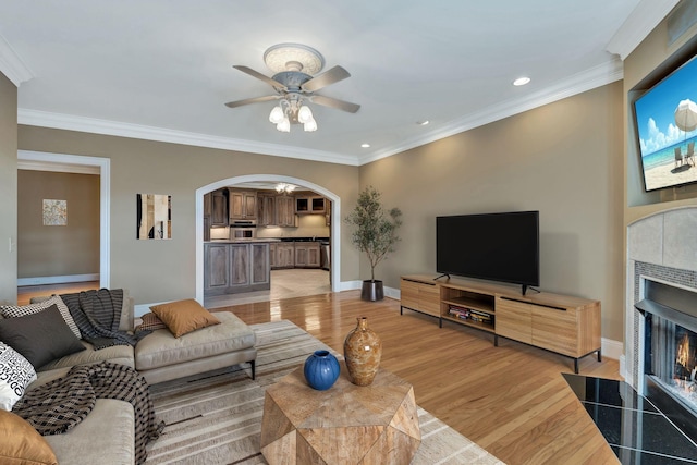 living area featuring baseboards, light wood-style flooring, arched walkways, a tiled fireplace, and crown molding