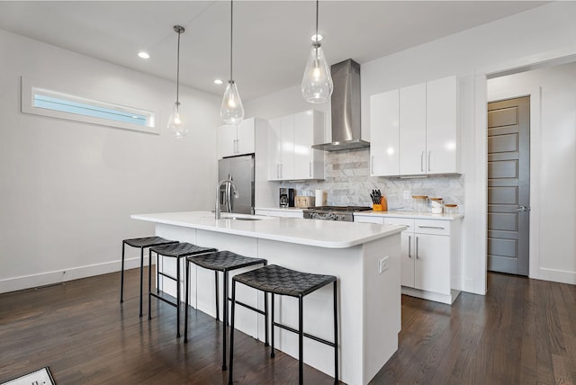 kitchen featuring an island with sink, a sink, backsplash, stainless steel fridge with ice dispenser, and wall chimney range hood