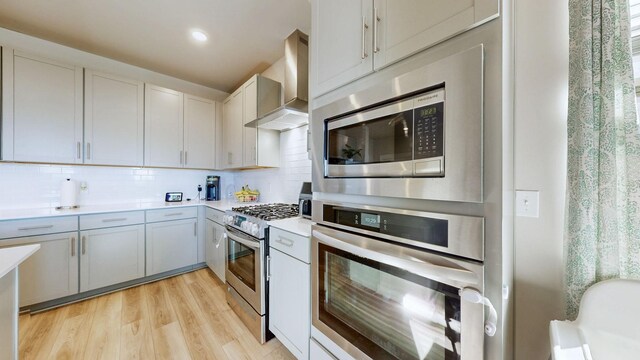 kitchen featuring light countertops, light wood-style floors, appliances with stainless steel finishes, wall chimney exhaust hood, and tasteful backsplash