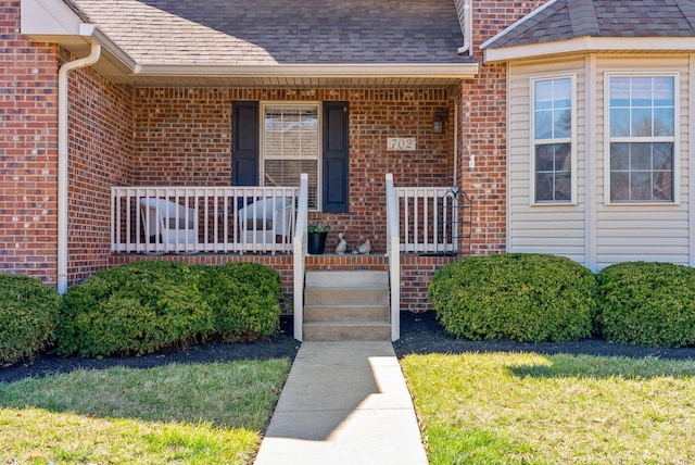 view of exterior entry with a porch, a yard, brick siding, and roof with shingles