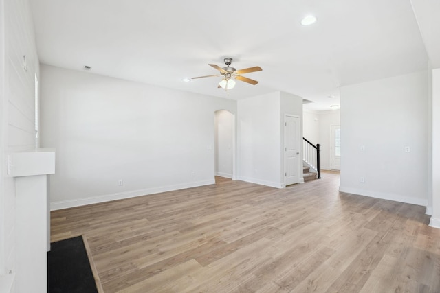 unfurnished living room featuring baseboards, stairway, light wood-type flooring, arched walkways, and a ceiling fan