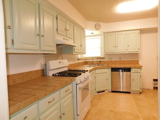 kitchen featuring tile countertops, gas range gas stove, a sink, under cabinet range hood, and stainless steel dishwasher