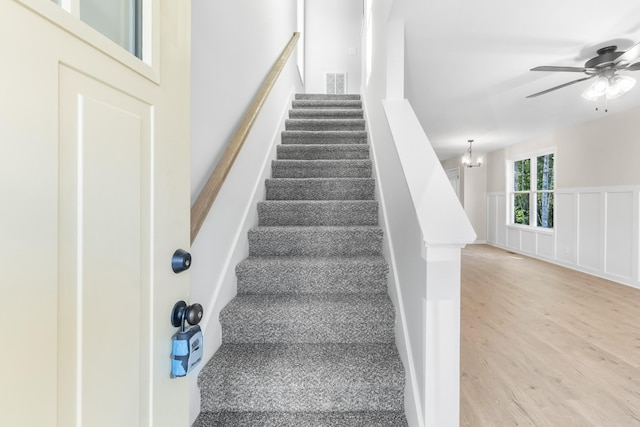 staircase featuring visible vents, a wainscoted wall, ceiling fan with notable chandelier, wood finished floors, and a decorative wall