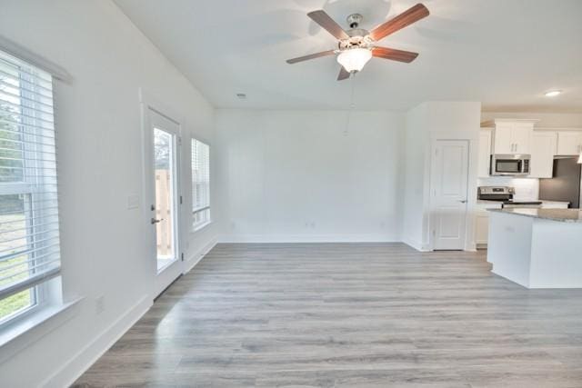 unfurnished living room featuring a wealth of natural light, light wood-style flooring, a ceiling fan, and baseboards