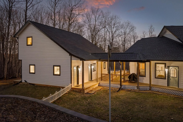 back of house at dusk with a yard, a shingled roof, a wooden deck, and fence