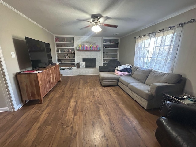 living room with built in shelves, a ceiling fan, a stone fireplace, dark wood-type flooring, and crown molding