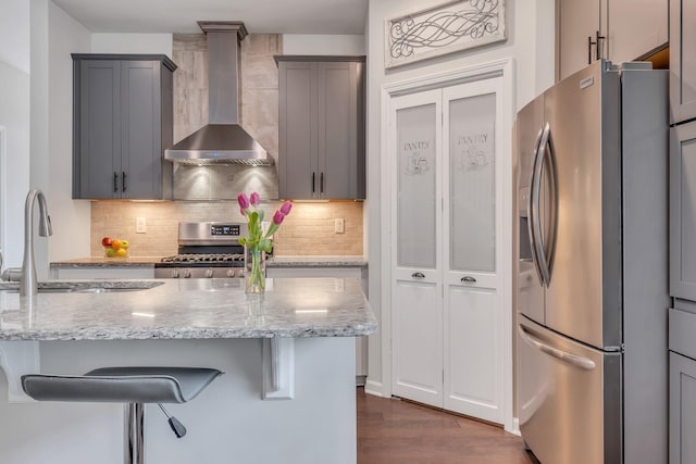 kitchen with wall chimney range hood, light stone countertops, a breakfast bar area, decorative backsplash, and appliances with stainless steel finishes