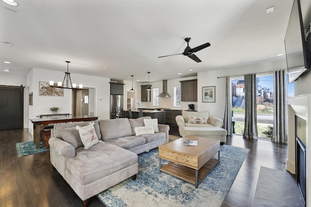 living room featuring a barn door, recessed lighting, ceiling fan with notable chandelier, and dark wood-style flooring