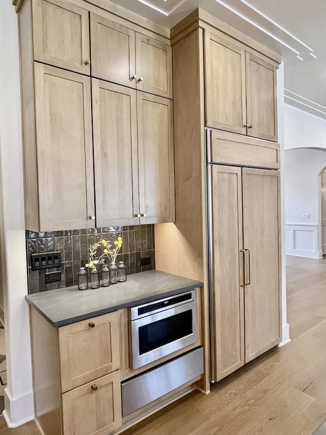 kitchen with tasteful backsplash, light brown cabinets, light wood-type flooring, paneled built in fridge, and arched walkways