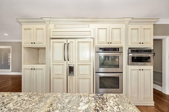 kitchen with ornamental molding, light stone counters, cream cabinets, stainless steel double oven, and paneled refrigerator