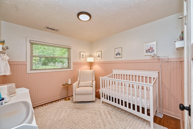 bedroom featuring visible vents, a crib, a textured ceiling, and a wainscoted wall