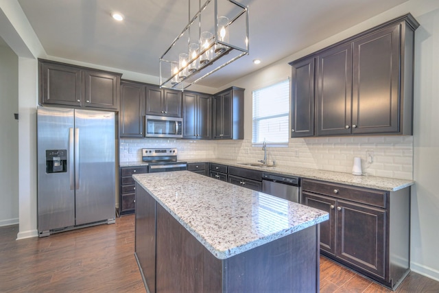 kitchen with a sink, a kitchen island, dark wood finished floors, appliances with stainless steel finishes, and light stone countertops