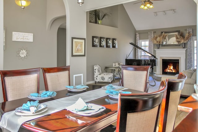 dining room featuring ceiling fan, vaulted ceiling, a warm lit fireplace, rail lighting, and arched walkways