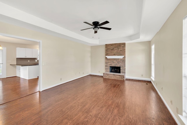 unfurnished living room with baseboards, a tray ceiling, a fireplace, ceiling fan, and dark wood-type flooring