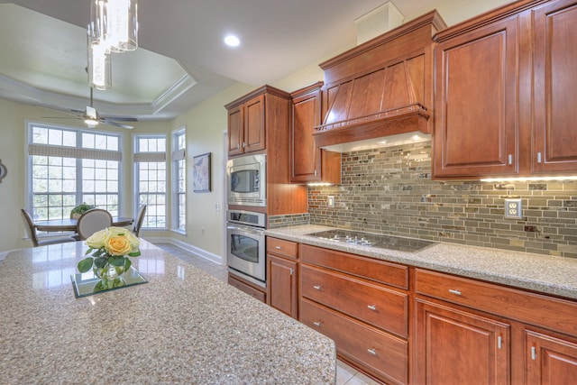 kitchen with brown cabinetry, stainless steel appliances, decorative backsplash, custom range hood, and a raised ceiling