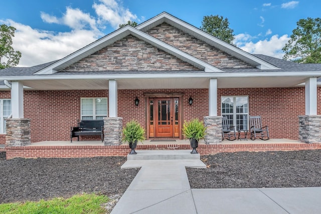 view of exterior entry featuring brick siding, stone siding, covered porch, and a shingled roof