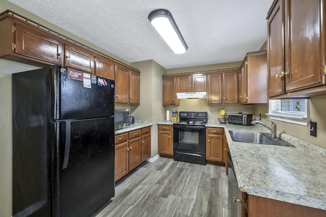 kitchen with under cabinet range hood, light wood-style flooring, brown cabinets, black appliances, and a sink