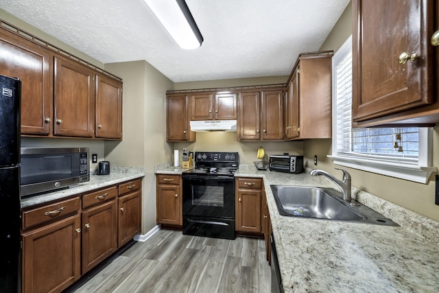 kitchen featuring a sink, black appliances, under cabinet range hood, a textured ceiling, and light wood-type flooring